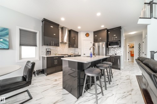 The kitchen features a center island with a sink and seating, glossy dark cabinetry, a stainless steel refrigerator, and a coordinating range hood - 3514 49 Avenue, Beaumont, AB - Indoor Photo Showing Other Room