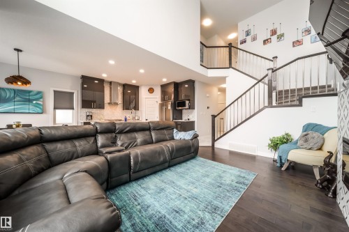 The property features an open-concept living space with dark hardwood flooring, a modern kitchen with dark cabinetry and stainless steel appliances, and a grand staircase with white risers and dark handrails - 3514 49 Avenue, Beaumont, AB - Indoor Photo Showing Living Room