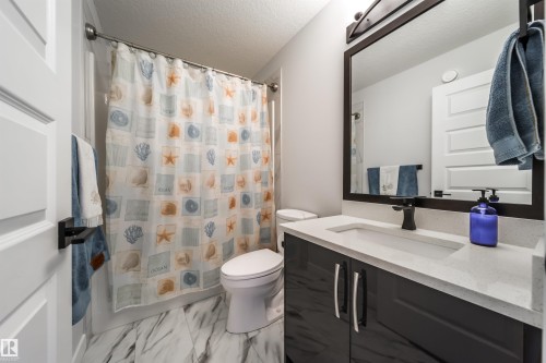 The bathroom features a vanity with a white countertop and dark cabinetry, a toilet, and a shower with a patterned curtain - 3514 49 Avenue, Beaumont, AB - Indoor Photo Showing Bathroom