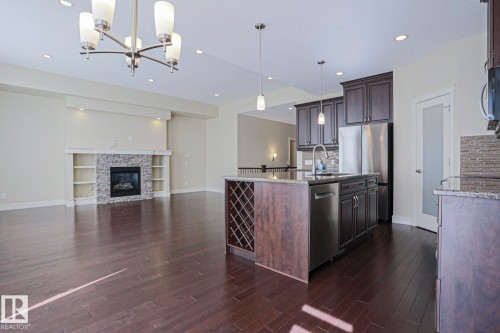 Expansive open-concept living space featuring hardwood floors, a stone-faced fireplace with built-in shelving, and a kitchen island with integrated wine storage - 2315 Ashcraft Cape, Edmonton, AB - Indoor Photo Showing Kitchen With Fireplace With Upgraded Kitchen