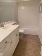 Bathroom featuring a white vanity with a white countertop and an integrated sink, a toilet, a bathtub with white tiled walls, and tiled flooring - 