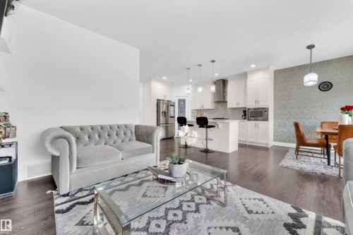 An open concept living area featuring dark wood flooring, a modern kitchen with white cabinetry and stainless steel appliances, and a dining area with patterned wallpaper - 2630 16A Ave Nw, Edmonton, AB - Indoor Photo Showing Living Room