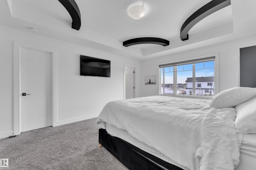 This room features a high ceiling with decorative black arch accents, a large window providing views of the neighborhood, and gray carpeting - 2630 16A Ave Nw, Edmonton, AB - Indoor Photo Showing Bedroom