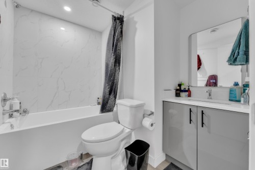 The bathroom features a bathtub with white tiled walls, a toilet, and a vanity with a white countertop and gray cabinets - 2630 16A Ave Nw, Edmonton, AB - Indoor Photo Showing Bathroom