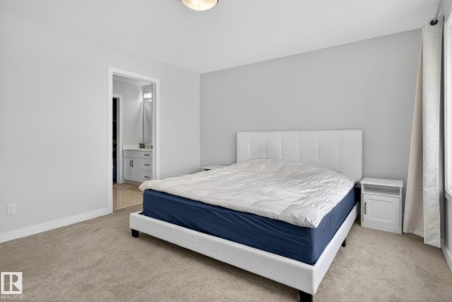 The bedroom features light-colored carpeting and walls, a window with drapery, and a visible doorway leading to a bathroom with a vanity - 3230 Parker Loop, Edmonton, AB - Indoor Photo Showing Bedroom