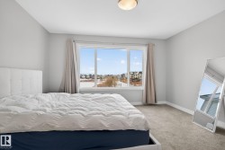 This room features light grey walls and carpet, with a large window providing ample natural light and views of the surrounding neighborhood - 
