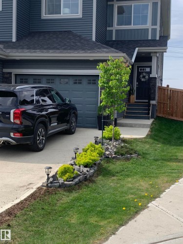 The property features a dark gray garage door, a dark gray exterior with stone accents, and a front yard with a well-maintained lawn and planted shrubs - 7490 Creighton Place, Edmonton, AB - Outdoor With Facade