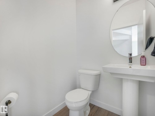 Bathroom featuring a white pedestal sink with a chrome faucet, a round mirror, and a white toilet - 95 1010 Rabbit Hill Road, Edmonton, AB - Indoor Photo Showing Bathroom