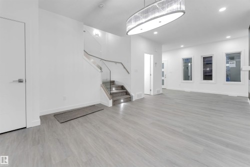 Bright open-concept living space featuring light-toned flooring, a modern chandelier, recessed lighting, and a contemporary staircase with glass railing - 444 Crystal Creek Link, Leduc, AB - Indoor Photo Showing Other Room