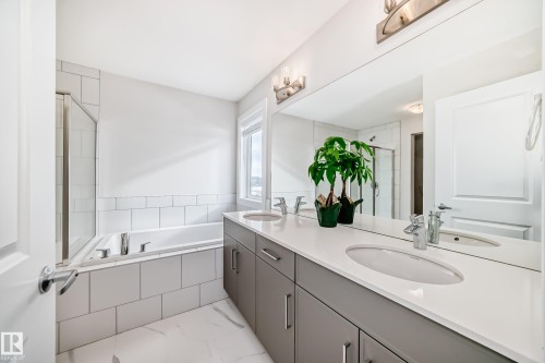Bathroom featuring a dual vanity with a white countertop and gray cabinetry, a built-in bathtub with white tile surround, a glass-enclosed shower, and a window providing natural light - 2935 Chokecherry Common, Edmonton, AB - Indoor Photo Showing Bathroom