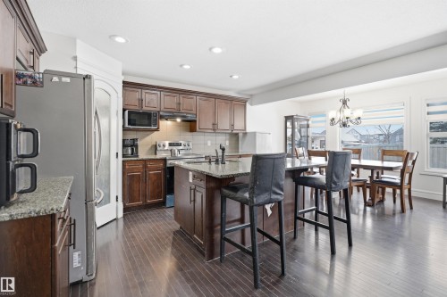 The kitchen features dark wood cabinetry, granite countertops, and stainless steel appliances, including a refrigerator, microwave, and stove - 469 Ainslie Crescent Sw, Edmonton, AB - Indoor Photo Showing Kitchen