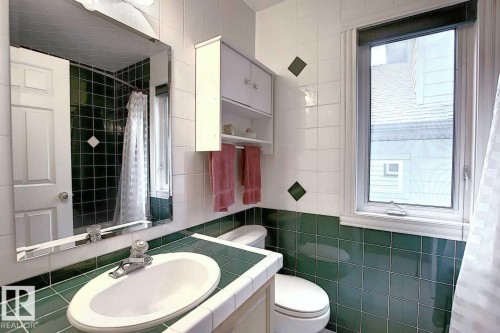 The bathroom features a window for natural light, white and green tiled walls, a vanity with a white countertop and oval sink, and a mirrored medicine cabinet - 11126 52 Street, Edmonton, AB - Indoor Photo Showing Bathroom