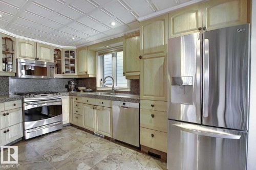 This kitchen features light wood cabinetry, stone countertops, and stainless steel appliances including a refrigerator, dishwasher, and stove with an overhead microwave - 11126 52 Street, Edmonton, AB - Indoor Photo Showing Kitchen With Upgraded Kitchen