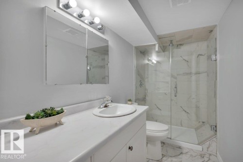 This bathroom features a vanity with a white countertop and an integrated sink, a wall-mounted mirrored medicine cabinet, and a shower with glass enclosure and marble-patterned tile - 12140 53 Street, Edmonton, AB - Indoor Photo Showing Bathroom