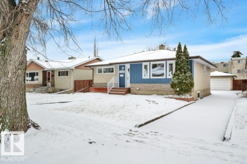 The property features a ranch-style exterior with blue siding and a beige facade, a front yard with a mature tree, and a driveway leading to a detached garage - 12140 53 Street, Edmonton, AB - Outdoor