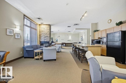Spacious common area featuring a stone accent fireplace, large windows with roller shades, and a kitchen area with wood cabinetry and a black refrigerator - 217 160 Magrath Road, Edmonton, AB - Indoor