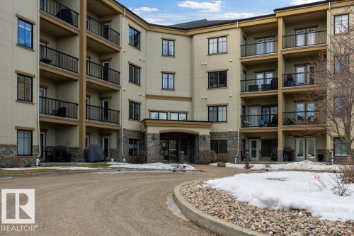 The property features a multi-story exterior with balconies, a paved circular driveway, and stone accents on the lower facade - 217 160 Magrath Road, Edmonton, AB - Outdoor With Balcony With Facade