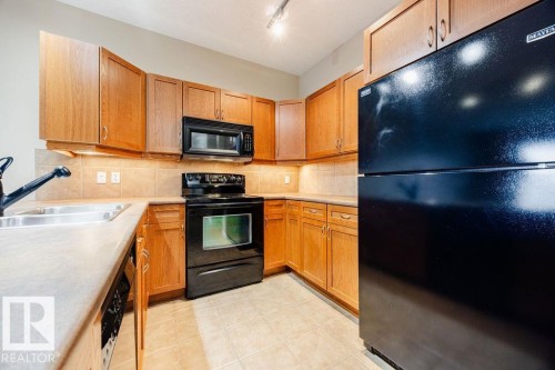 Kitchen featuring lots of wood cabinetry, a peninsula providing loads of counter top space, tile backsplash, a stainless steel sink, black appliances and tiled flooring - 217 160 Magrath Road, Edmonton, AB - Indoor Photo Showing Kitchen With Double Sink