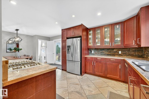 The kitchen features extensive wood cabinetry with some upper cabinets having glass fronts, a stainless steel refrigerator, and a stone-tiled backsplash - 15701 92A Avenue, Edmonton, AB - Indoor Photo Showing Kitchen