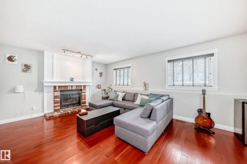 This inviting living space features gleaming hardwood floors, a prominent brick fireplace with a white mantel, and two windows providing natural light - 3107 43 Avenue, Edmonton, AB - Indoor Photo Showing Living Room With Fireplace