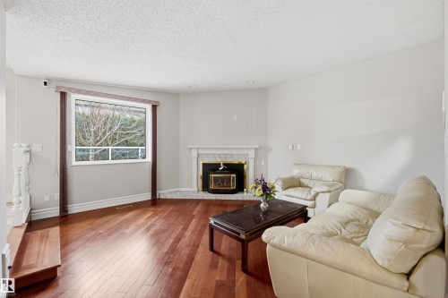 Living room featuring rich hardwood floors, a corner fireplace with a white mantel and marble surround, and a large window providing natural light - 131 Walsh Crescent, Edmonton, AB - Indoor Photo Showing Living Room With Fireplace