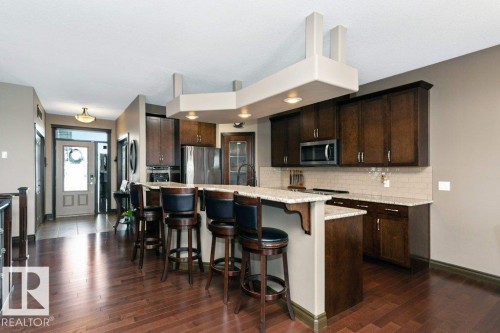 The kitchen features dark wood cabinetry, stainless steel appliances, a white tile backsplash, and a granite island with seating - 33 Hillcrest Point(E), Fort Saskatchewan, AB - Indoor Photo Showing Kitchen With Stainless Steel Kitchen With Upgraded Kitchen