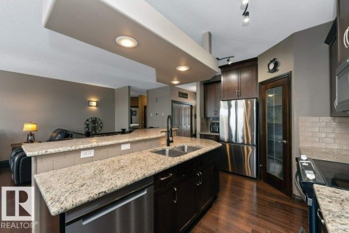The kitchen features granite countertops, dark wood cabinetry, and stainless steel appliances, including a side-by-side refrigerator and dishwasher - 33 Hillcrest Point(E), Fort Saskatchewan, AB - Indoor Photo Showing Kitchen With Stainless Steel Kitchen With Double Sink With Upgraded Kitchen
