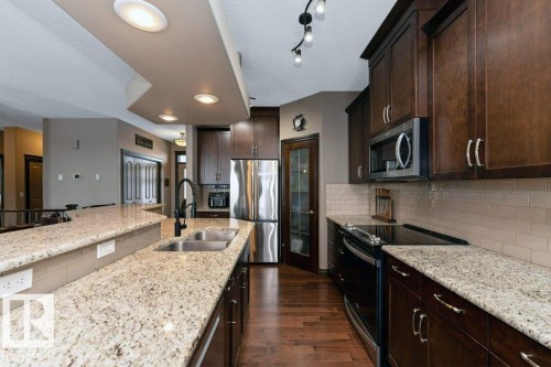 Well-appointed kitchen featuring granite countertops, dark wood cabinetry, stainless steel appliances, a tile backsplash, and recessed lighting - 33 Hillcrest Point(E), Fort Saskatchewan, AB - Indoor Photo Showing Kitchen With Stainless Steel Kitchen With Double Sink With Upgraded Kitchen