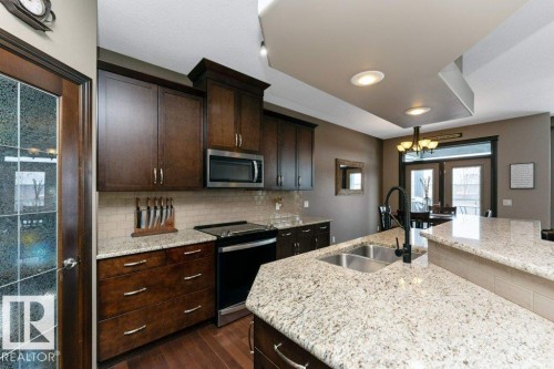 The kitchen features wood cabinetry, a stainless steel microwave, a black stove, light-colored granite countertops, and a double basin sink with a black faucet - 33 Hillcrest Point(E), Fort Saskatchewan, AB - Indoor Photo Showing Kitchen With Double Sink