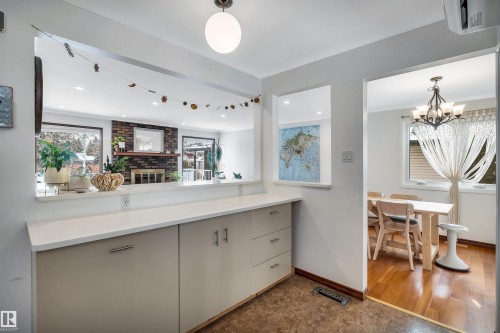This interior view showcases a built-in counter with light-colored cabinetry and a white countertop, offering a pass-through opening to a living area with a brick fireplace and windows - 4208 89 Street Nw, Edmonton, AB - Indoor