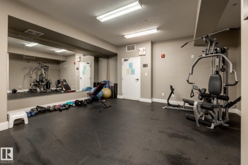 The property offers a fitness room with rubber flooring, a large wall-mounted mirror, and overhead lighting - 454 6079 Maynard Way, Edmonton, AB - Indoor Photo Showing Gym Room