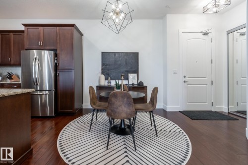 This open-concept dining area features rich hardwood floors, a contemporary geometric light fixture, and a built-in kitchen with dark wood cabinetry and a stainless steel refrigerator - 454 6079 Maynard Way, Edmonton, AB - Indoor