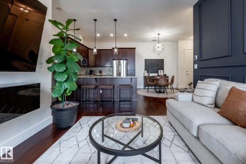 The property offers an open concept layout with dark hardwood flooring, a kitchen featuring dark wood cabinetry and a breakfast bar, and a dining area with a modern chandelier - 454 6079 Maynard Way, Edmonton, AB - Indoor Photo Showing Living Room With Fireplace