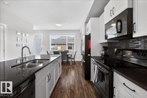 The kitchen features an island with a double basin sink and a modern faucet, dark countertops, white cabinetry, and black appliances - 9034 Rosenthal Link Link, Edmonton, AB - Indoor Photo Showing Kitchen With Double Sink With Upgraded Kitchen