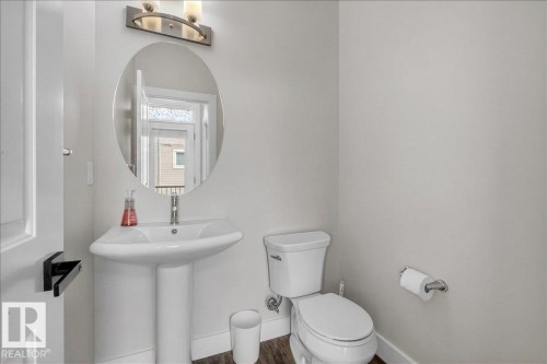This bathroom features a white pedestal sink, an oval mirror, a modern vanity light fixture, and a toilet with a chrome toilet paper holder - 9034 Rosenthal Link Link, Edmonton, AB - Indoor Photo Showing Bathroom