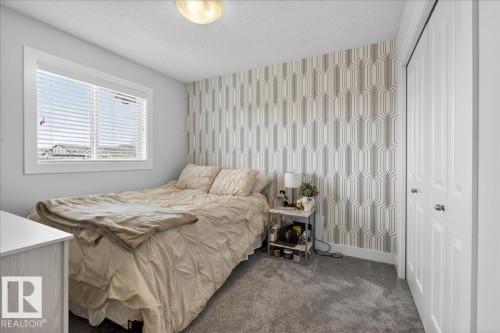 This room features a window with horizontal blinds, a neutral-toned patterned accent wall, and soft grey carpeting - 9034 Rosenthal Link Link, Edmonton, AB - Indoor Photo Showing Bedroom