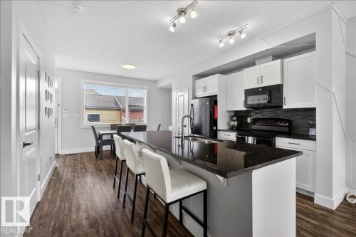 The kitchen features white cabinetry, a black tile backsplash, and a black granite island with a sink - 9034 Rosenthal Link Link, Edmonton, AB - Indoor Photo Showing Kitchen With Upgraded Kitchen