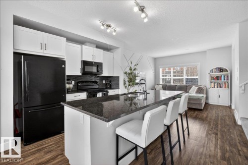 The kitchen features white cabinetry, a large kitchen island with a dark countertop and seating for three, and black appliances including a refrigerator, oven, and microwave - 9034 Rosenthal Link Link, Edmonton, AB - Indoor Photo Showing Kitchen