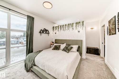 This bedroom features a large window with views of the surrounding neighborhood, light-colored carpeting, and a ceiling light fixture - 10936 117 Street, Edmonton, AB - Indoor Photo Showing Bedroom