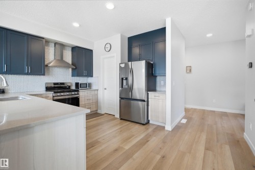 The kitchen features dark blue and light wood cabinetry, light countertops, stainless steel appliances, and a white tiled backsplash - 7017 54 Avenue, Beaumont, AB - Indoor Photo Showing Kitchen With Stainless Steel Kitchen With Upgraded Kitchen