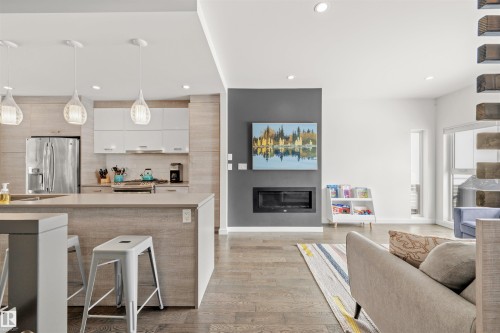 The open-concept living area features hardwood floors, a contemporary kitchen with light wood-grain cabinetry, and a living room with a modern fireplace and a large window - 10607 127 Street, Edmonton, AB - Indoor With Fireplace