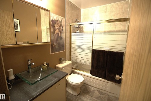 The bathroom features a glass vessel sink, a vanity with a dark countertop and light-colored cabinetry, a toilet, and a bathtub with a shower and sliding glass doors - 9347 175 Avenue, Edmonton, AB - Indoor Photo Showing Bathroom