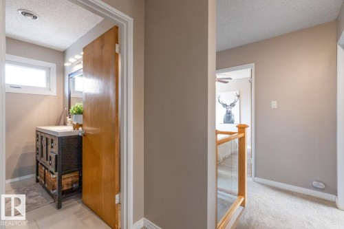 This interior view highlights a bathroom with a window and vanity, and a hallway featuring carpeting and a wooden banister with glass panels - 10504 60 Street, Edmonton, AB - Indoor Photo Showing Other Room