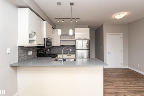 Kitchen featuring a spacious island with a sink and pendant lighting, stainless steel appliances, white cabinetry, and gray subway tile backsplash - 313 10418 81 Avenue, Edmonton, AB - Indoor Photo Showing Kitchen With Double Sink
