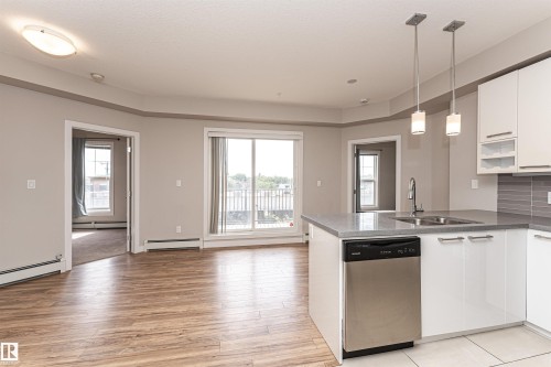 Open concept living area with wood-look flooring, a sliding glass door leading to a balcony, and a kitchen island featuring a stainless steel dishwasher and dual pendant lighting - 313 10418 81 Avenue, Edmonton, AB - Indoor Photo Showing Kitchen With Double Sink