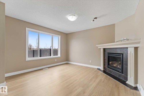 The living area features light-toned flooring, a large window providing natural light, and a fireplace with a stone-tile surround and mantel - 84 Calvert Wynd, Fort Saskatchewan, AB - Indoor Photo Showing Living Room With Fireplace