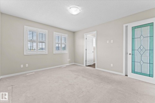 This room features light-colored carpet, two windows with white louvered shutters, and a decorative door with clear and teal patterned glass - 84 Calvert Wynd, Fort Saskatchewan, AB - Indoor Photo Showing Other Room