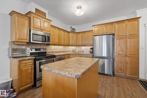 The kitchen features extensive wood cabinetry, a stainless steel refrigerator, and a kitchen island with a stone-look countertop - 318 9820 165 Street, Edmonton, AB - Indoor Photo Showing Kitchen