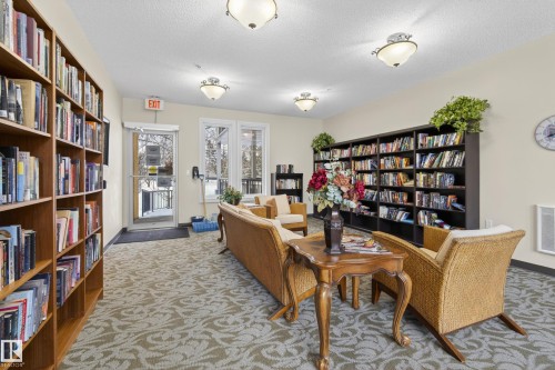 Inviting common area featuring extensive bookshelves, windows providing natural light, and patterned carpeting - 318 9820 165 Street, Edmonton, AB - Indoor Photo Showing Other Room