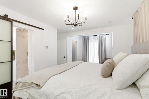 This bedroom features a light color palette, a modern chandelier, and sliding barn door access to a bathroom with a tiled shower - 38 Woodcrest Avenue, St. Albert, AB - Indoor Photo Showing Bedroom